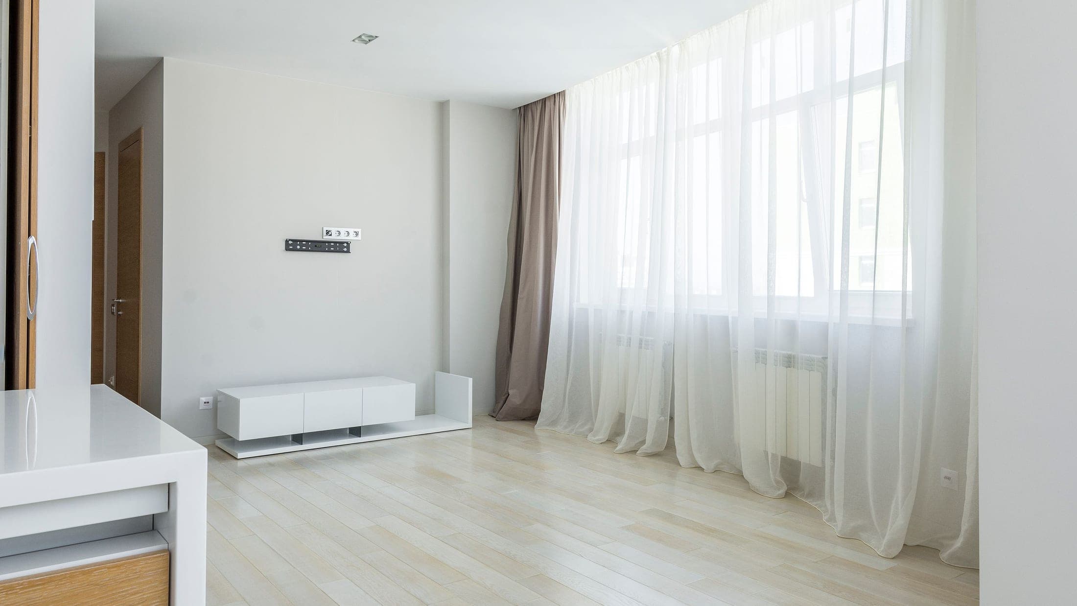 Séjour vide et lumineux au parquet clair, grandes fenêtres aux voilages blancs