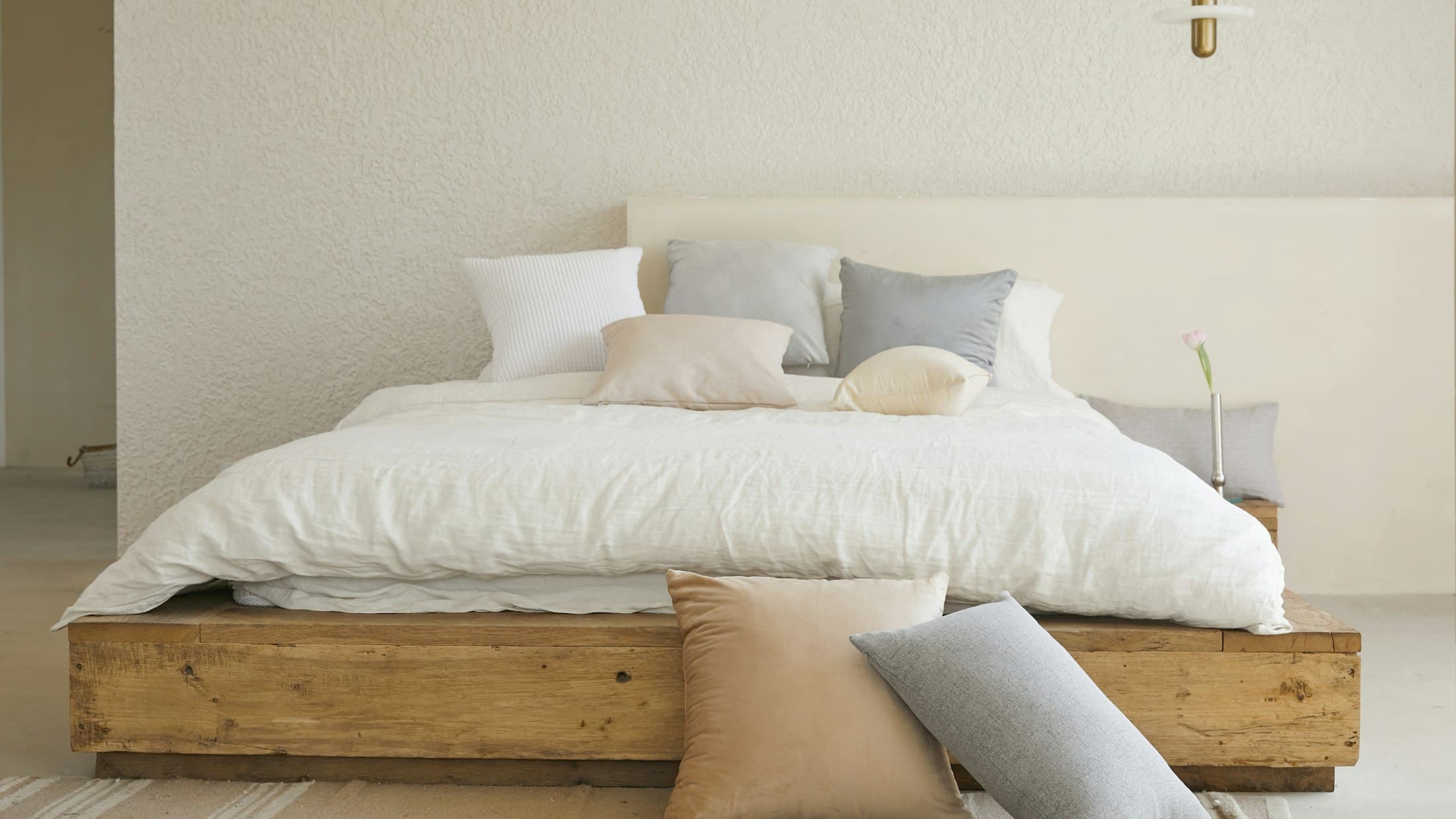 Lit fait sur un sommier en bois brut, couette blanche et coussins gris et beige, dans une chambre claire