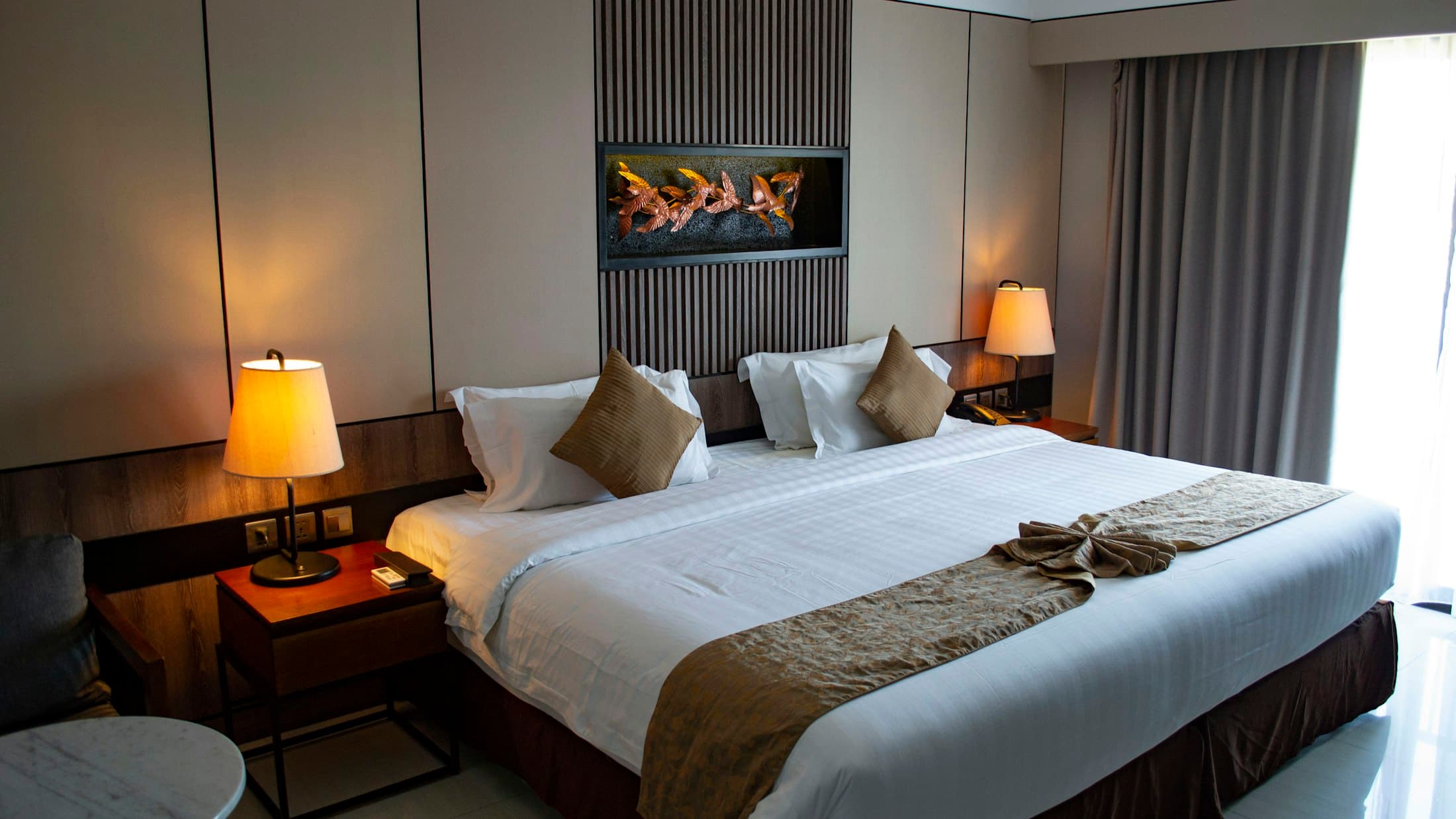 Chambre d'hôtel prête : grand lit aux draps blancs, chemin de lit brun noué, lampes de chevet allumées