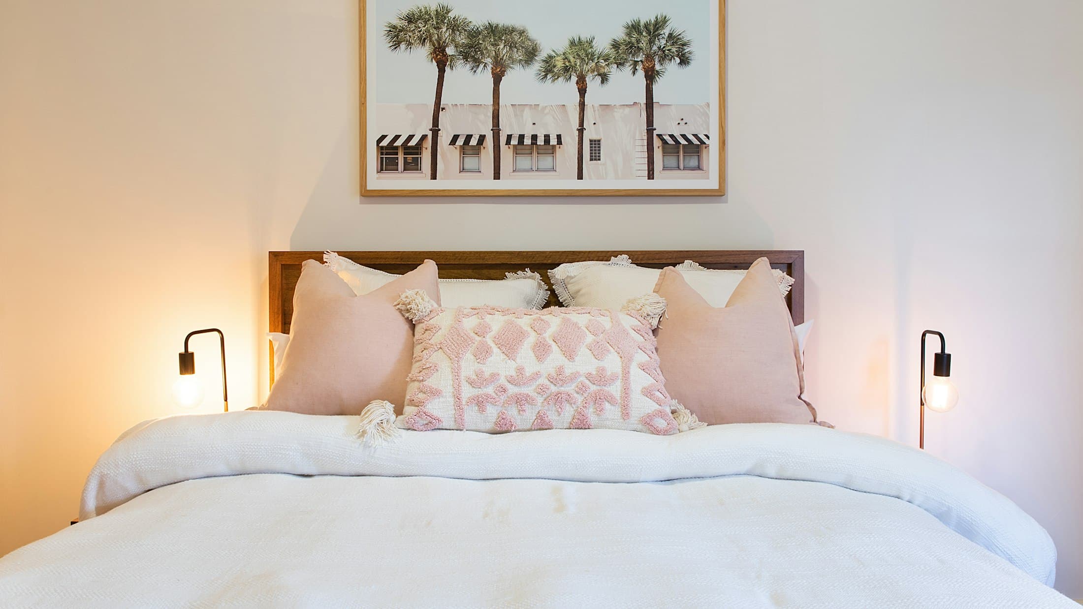 Chambre de location prête à accueillir : lit à la couette blanche, coussins roses, affiche de palmiers au mur